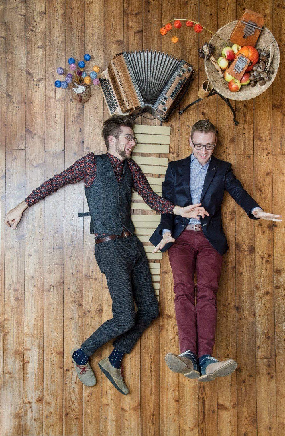 Jonny Kölbl und Leonhard Waltersdorfer liegen lachend auf einem Holzboden, umgeben von Steirischer Harmonika, Vibraphon-Stäben, Mbira und Äpfeln.