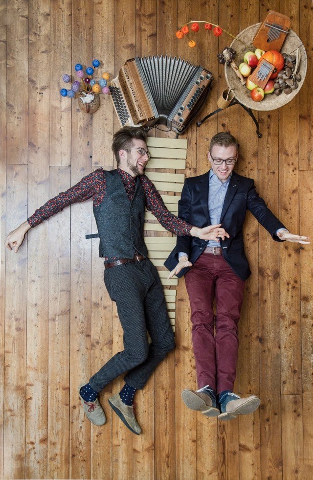 Duo press photo: Jonny Kölbl and Leonhard Waltersdorfer lying on a wooden floor, laughing, among accordion, vibraphone bars and mbira.