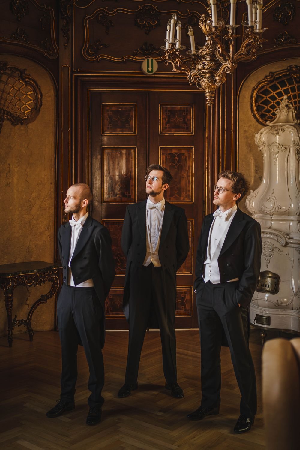 Jonny Kölbl, Leonhard Waltersdorfer und Hannes Schöggl im Frack vor einer holzvertäfelten Tür im Palais Auersperg, über ihnen ein Kronleuchter.