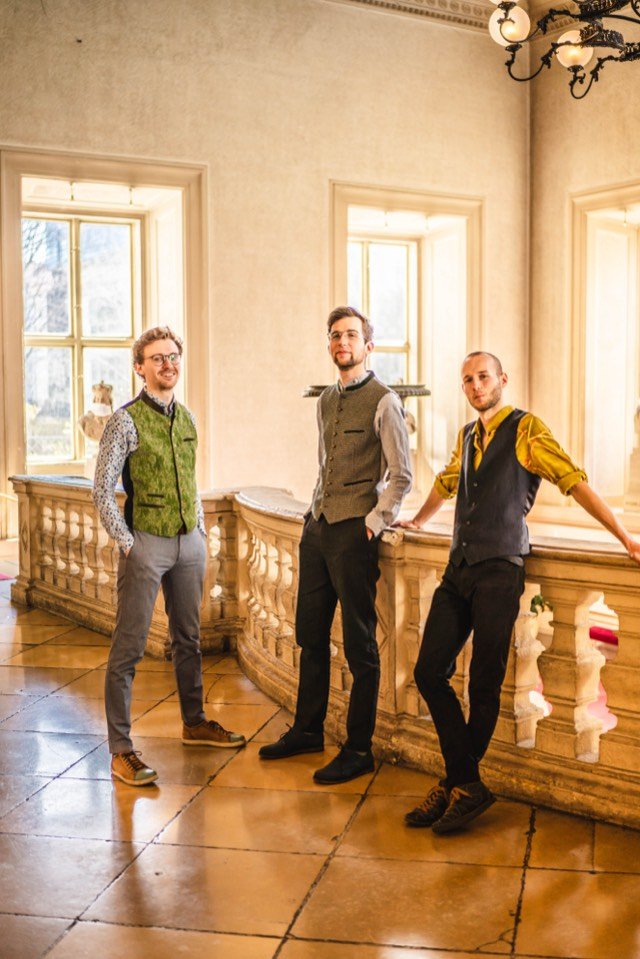 Die drei Musiker in Westen an einer Balustrade im lichtdurchfluteten Palais.
